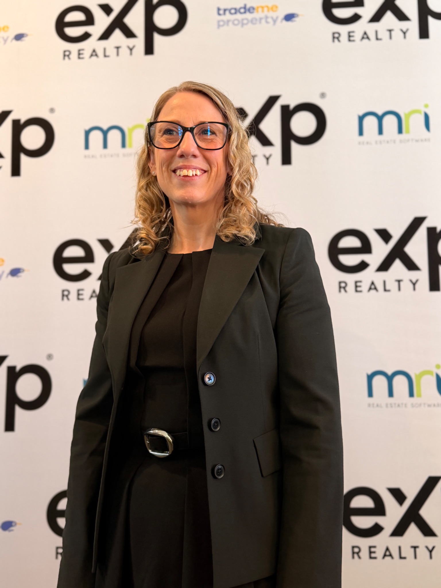 Jasmine Rohland in a black blazer in front of an eXp Realty branded backdrop