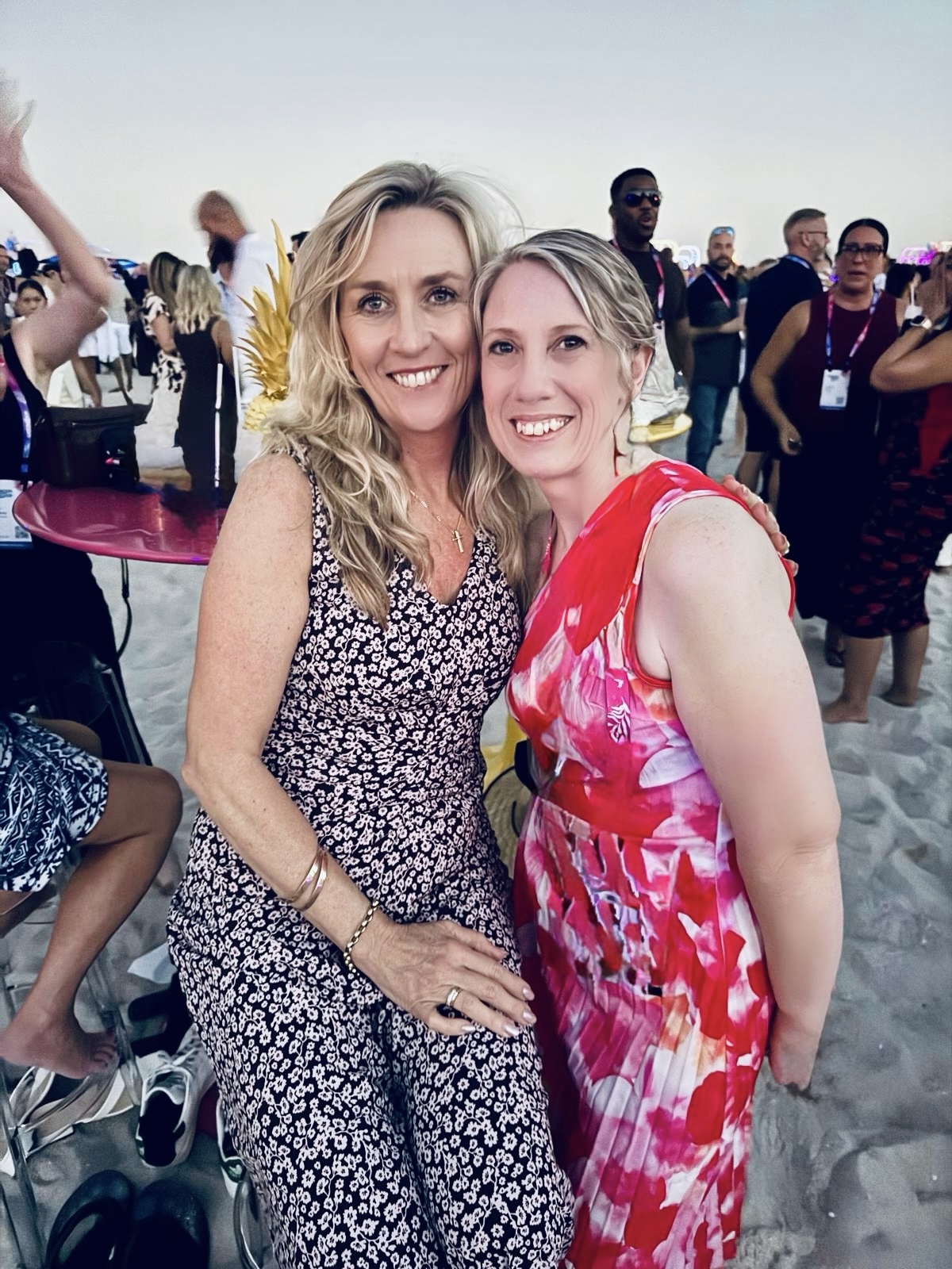 Two women smiling together at a gathering with other attendees in the background
